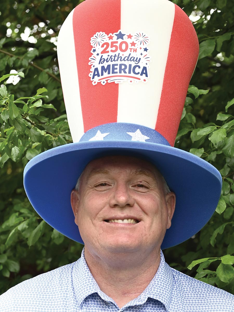 Brady L. Kay wears a tall patriotic hat, smiling against green leaves.
