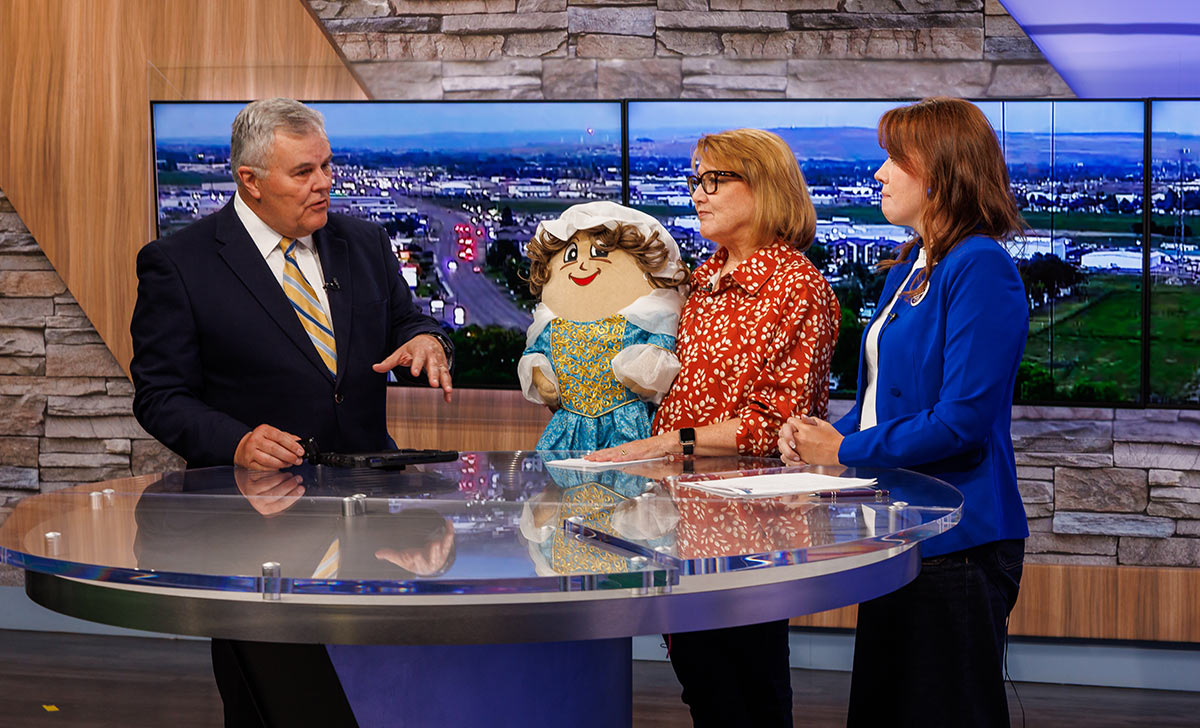 People on a television news set talking next to a large doll character.