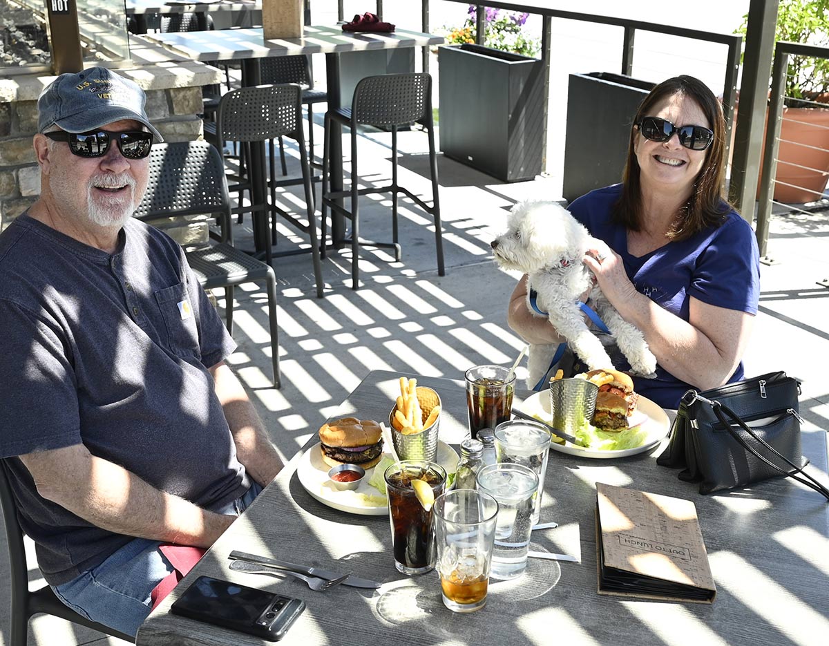 A couple smiles while dining on an outdoor patio with their small white dog and burgers.