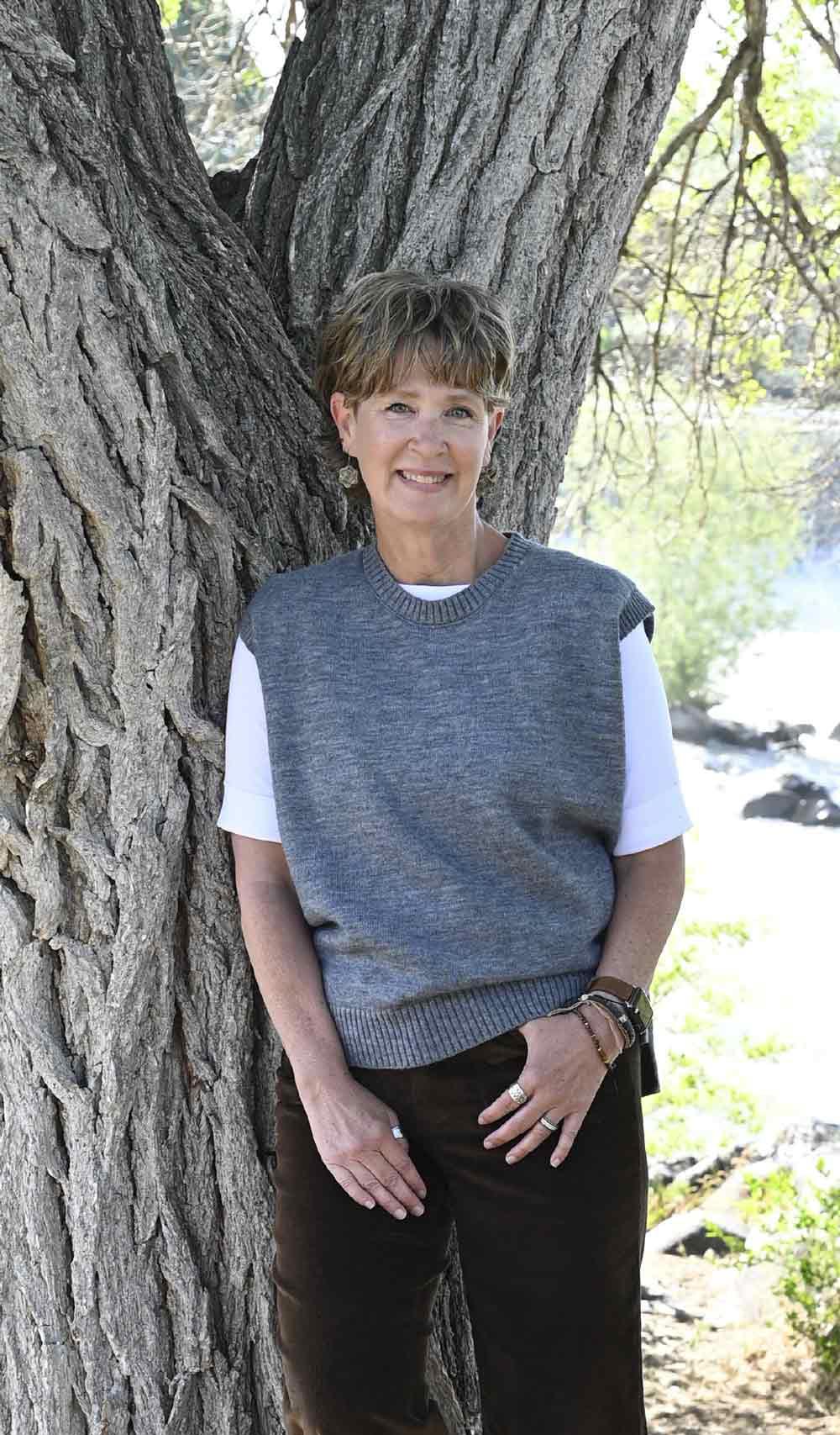 A smiling woman in a grey sweater vest and brown pants leans against a large tree trunk outdoors.