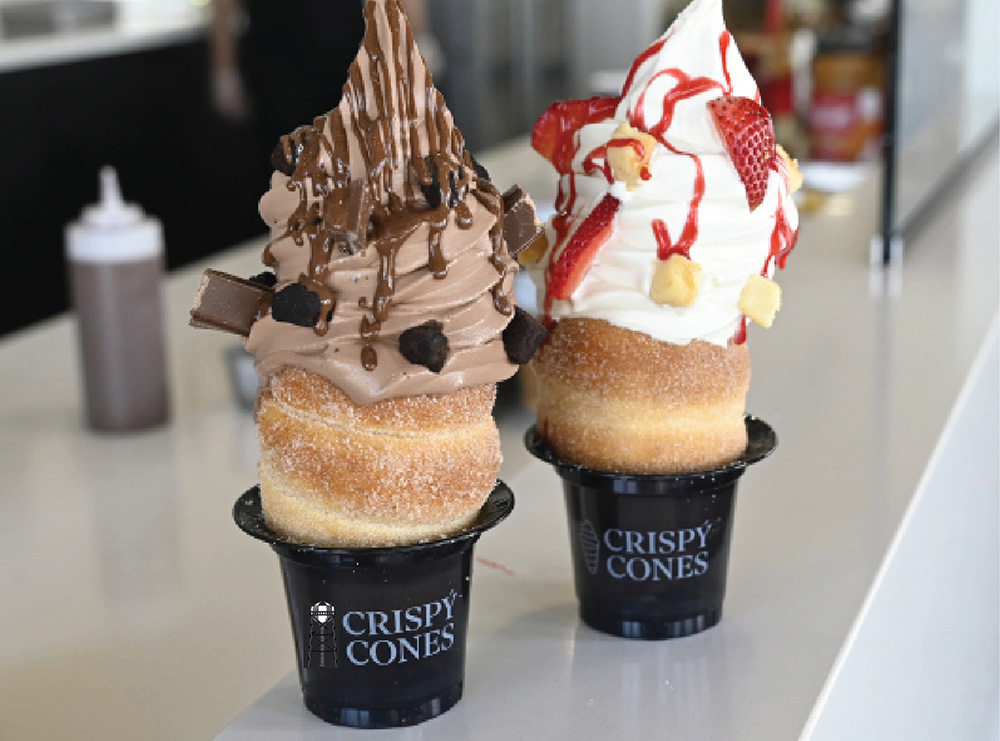 Two soft-serve ice cream treats served in sugar-dusted pastry cones—known as chimney cakes—which rest inside black plastic cups labeled CRISPY CONES