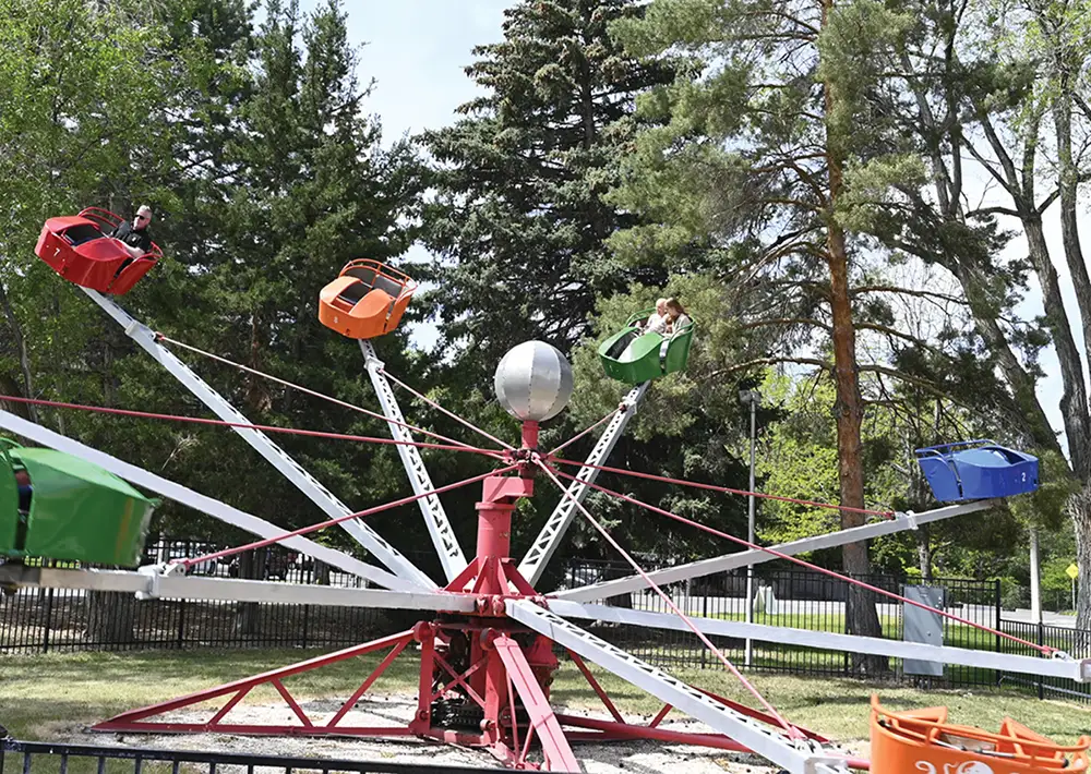 A spinning amusement park ride with a red central base, a silver sphere on top, and white metal arms carrying people in colorful green, red, orange, and blue cars, surrounded by tall green trees.