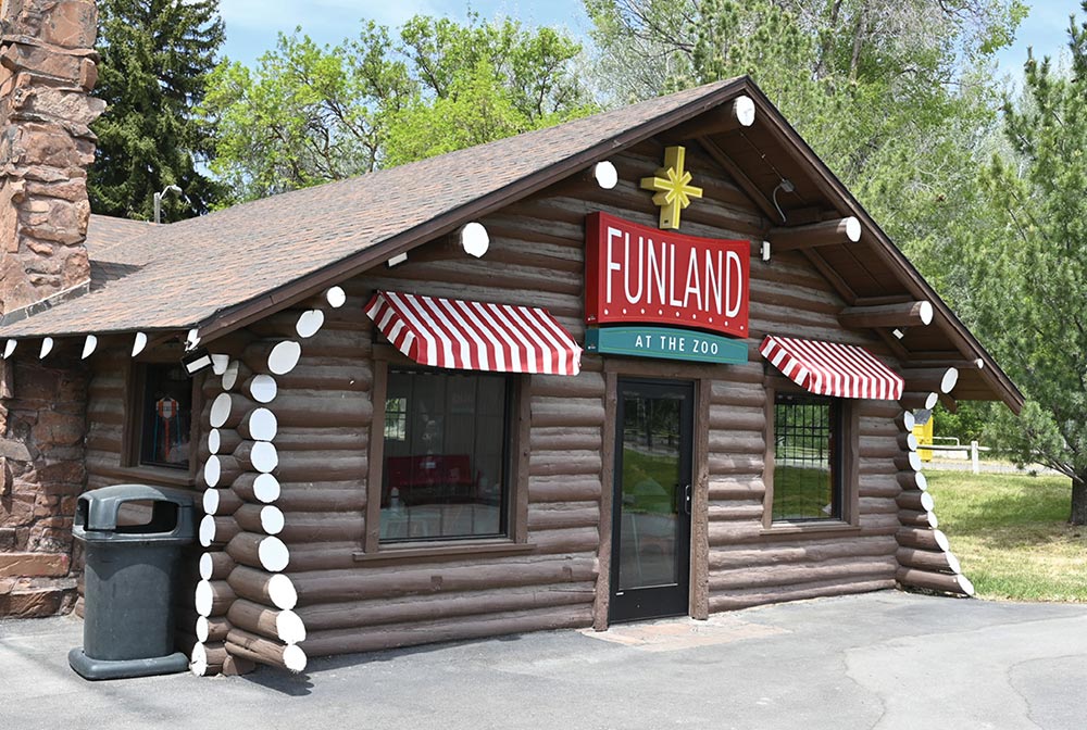 The exterior of a brown log cabin building featuring a stone chimney, red-and-white striped awnings over the windows, and a prominent red and green sign that reads "FUNLAND AT THE ZOO."
