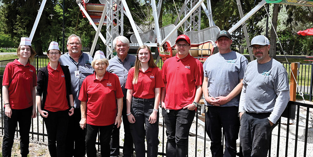 Nine people stand in a row posing for a group photo outdoors in front of a white metal Ferris wheel. Several individuals wear matching red polo shirts and white paper hats.