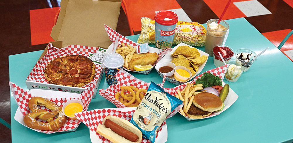 A turquoise table filled with amusement park food, including a pepperoni pizza in a box, a hot dog, a soft pretzel with cheese, a burger, fries, onion rings, fried fish, bags of popcorn, a bag of chips, and drinks.