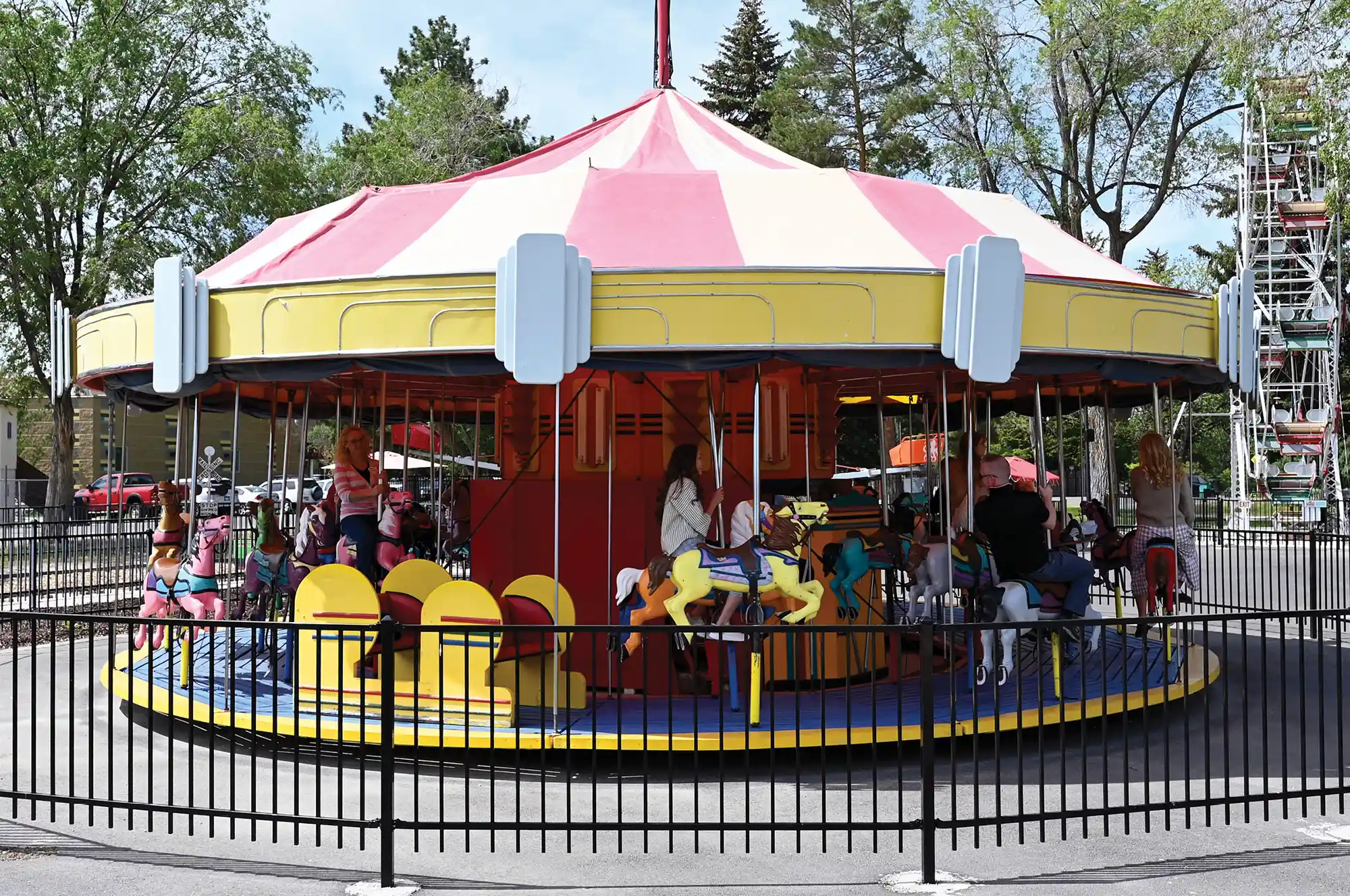 A carousel ride with a pink and white striped canopy and yellow trim. People sit on colorful horse figures on the rotating platform. A black metal fence and tall green trees surround the ride.