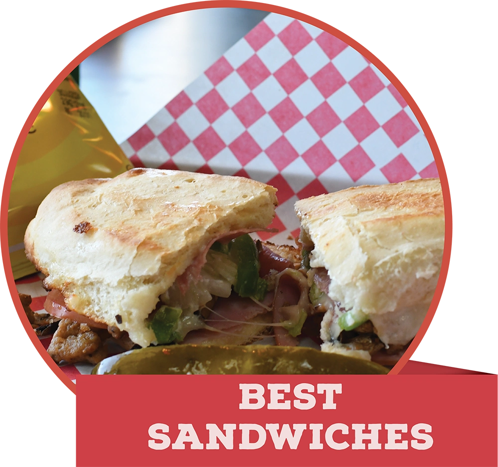 A toasted panini sandwich cut in half, served with a pickle spear, featuring a red banner reading "BEST SANDWICHES."