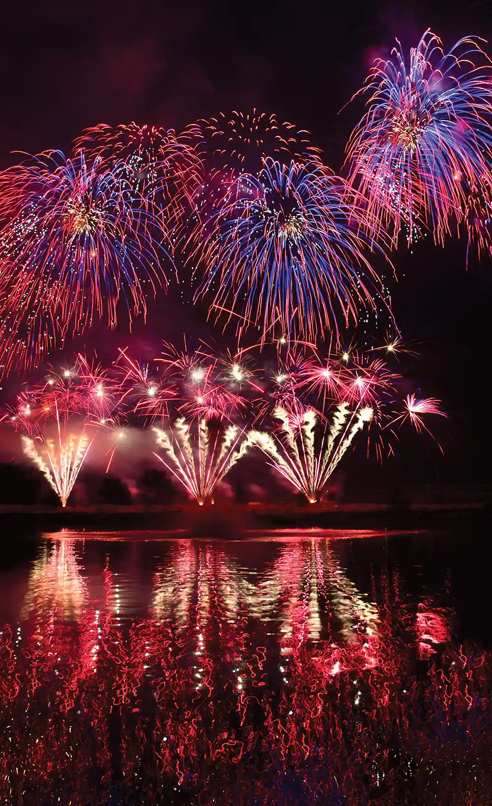 A vibrant nighttime display of red, blue, and gold fireworks exploding over water. Large spherical bursts fill the dark sky above lower fan-shaped flares, casting dazzling, colorful reflections on the rippling surface below.