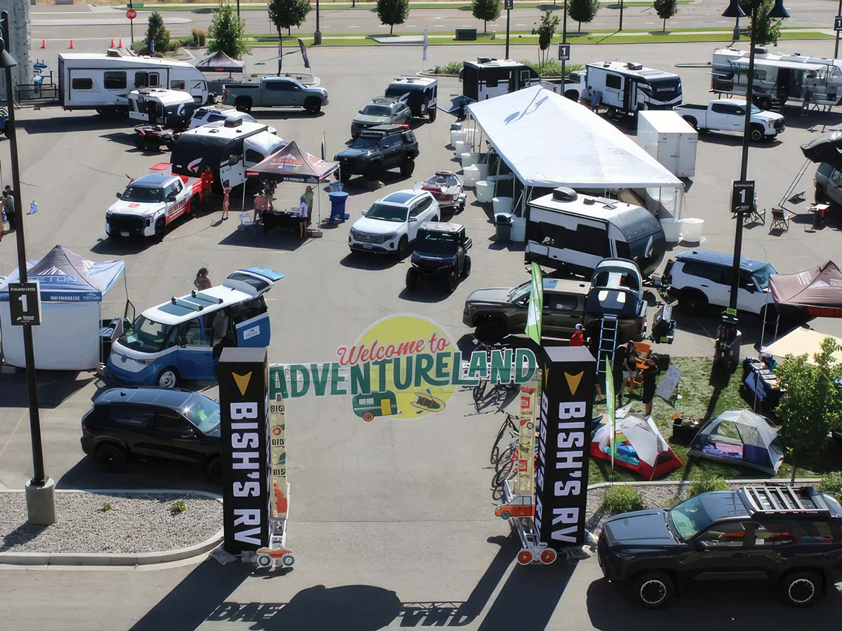 An elevated view of an outdoor RV expo in a parking lot, featuring an entrance arch reading "Welcome to Adventureland" by Bish's RV. The lot is filled with various recreational vehicles, camper vans, trucks, and vendor tents.