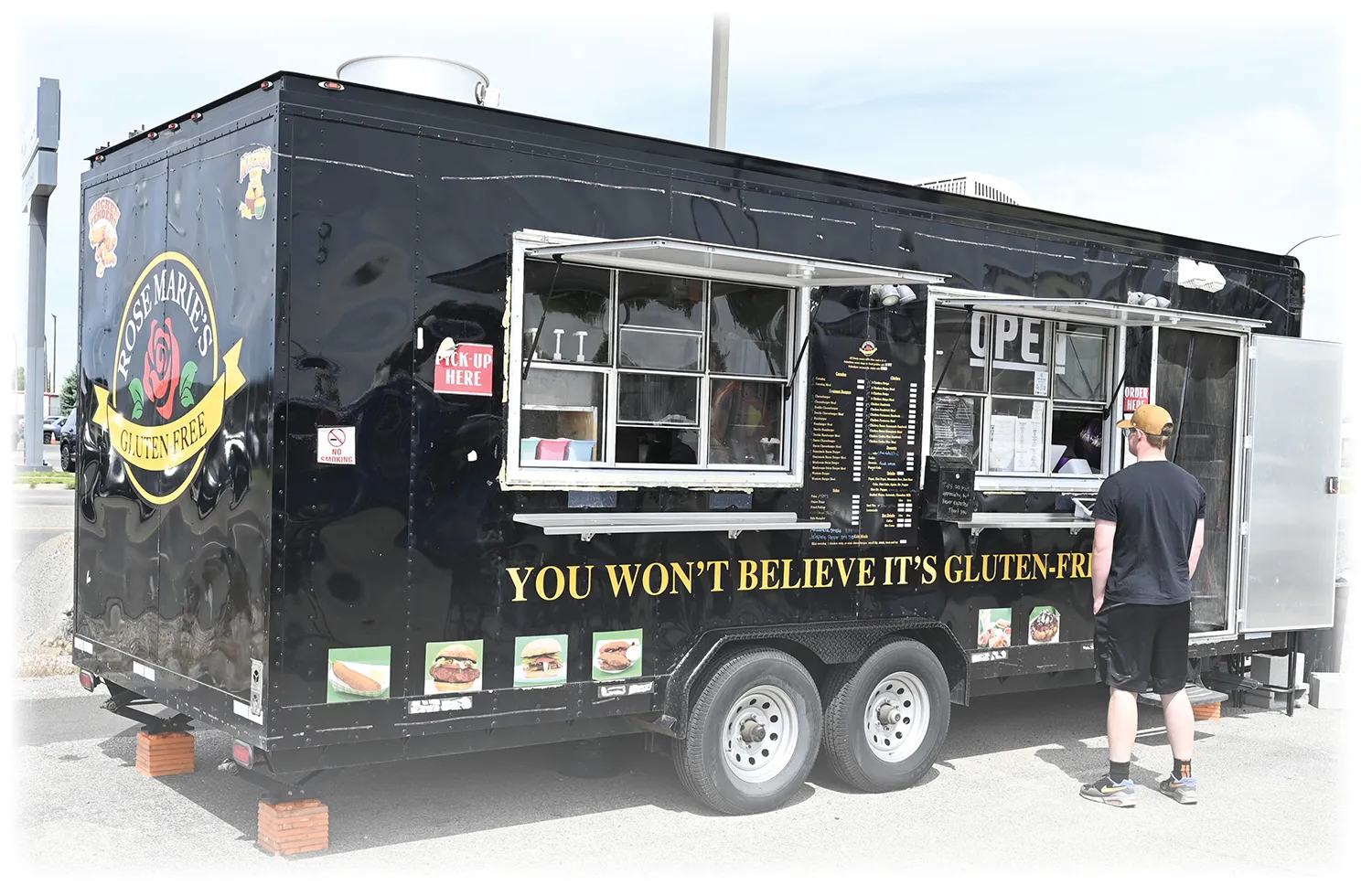 A black food trailer named "Rose Marie's Gluten Free" with the yellow slogan "YOU WON'T BELIEVE IT'S GLUTEN-FREE" painted on its side. A customer wearing a cap and shorts stands at the open service window.