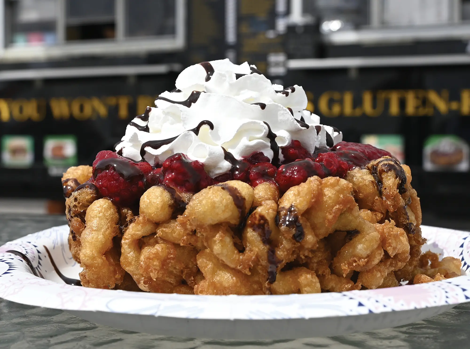 A golden-brown funnel cake on a paper plate, piled high with berries, whipped cream, and chocolate syrup, with a gluten-free food truck in the blurred background.