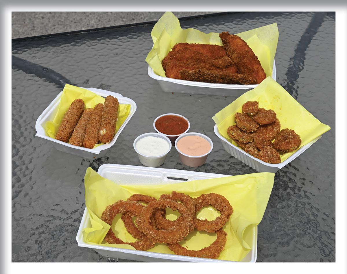 Four foam food containers sit on a glass table. Inside on yellow paper are breaded and fried foods: chicken tenders, mozzarella sticks, fried pickles, and onion rings. Three small plastic cups containing dipping sauces sit in the center.