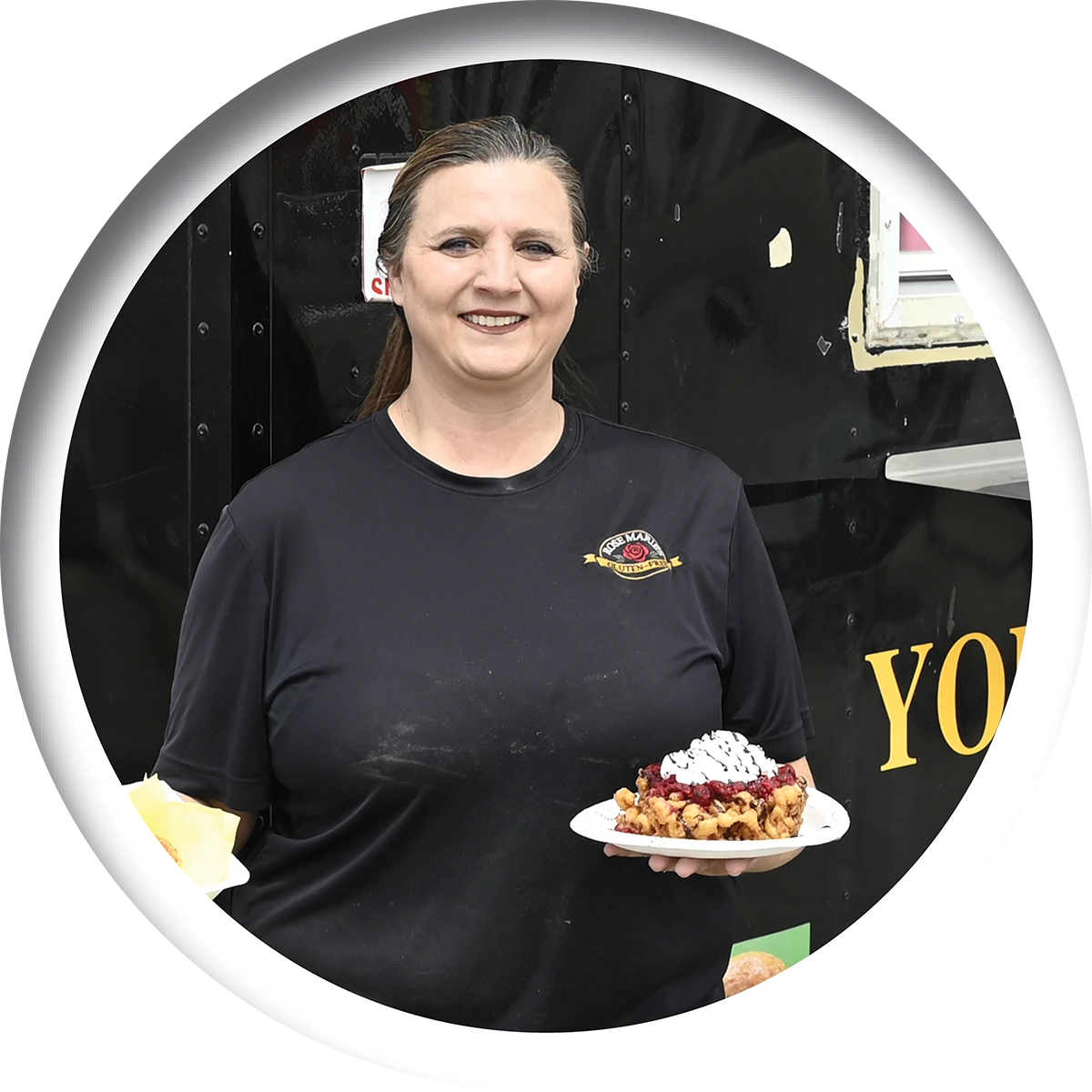 A smiling woman wearing a black Rose Marie's shirt holds a plate with a loaded funnel cake in front of a black food truck, shown within a circular crop.