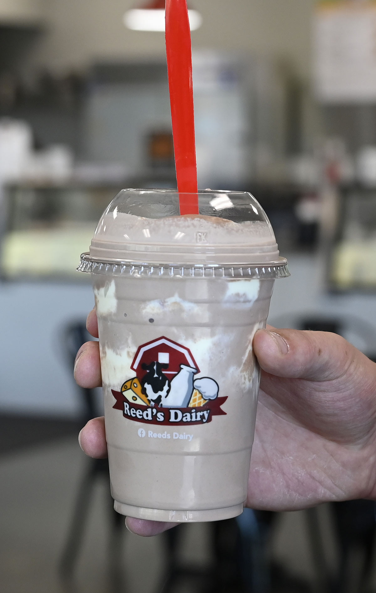 A hand holding a chocolate milkshake in a clear plastic cup featuring the Reed's Dairy logo, topped with a dome lid and a red spoon.