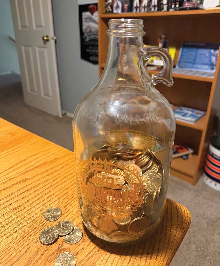 A glass half-gallon jug filled with presidential $1 coins sitting on a wooden table.