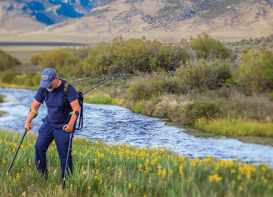 A man using forearm crutches walks along a scenic riverbank with a fishing rod.
