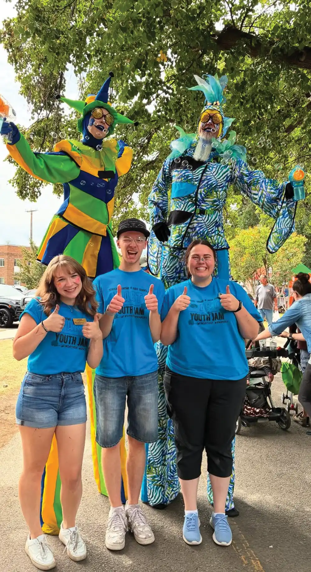 Three people in blue Youth Jam shirts giving thumbs up in front of two costumed stilt walkers blowing bubbles.