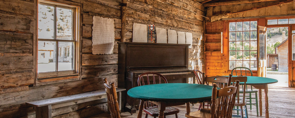 Interior of a rustic log cabin featuring wooden tables with green felt tops, chairs, and an upright piano.