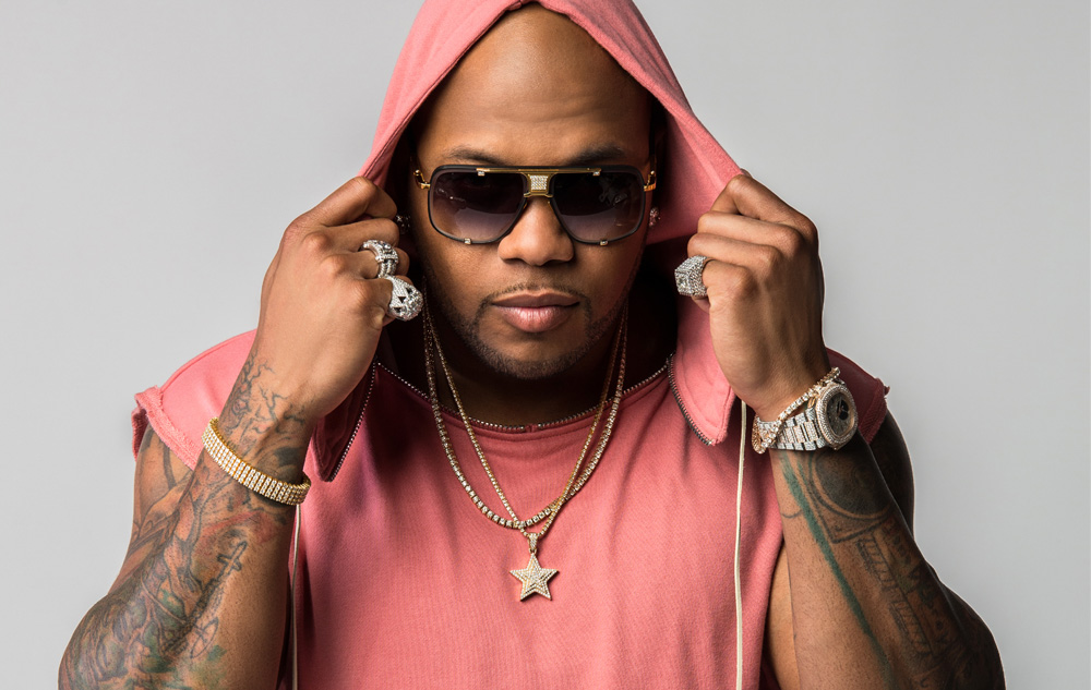 Rapper Flo Rida wearing a pink hoodie, sunglasses, and gold jewelry, adjusting his hood for the camera.