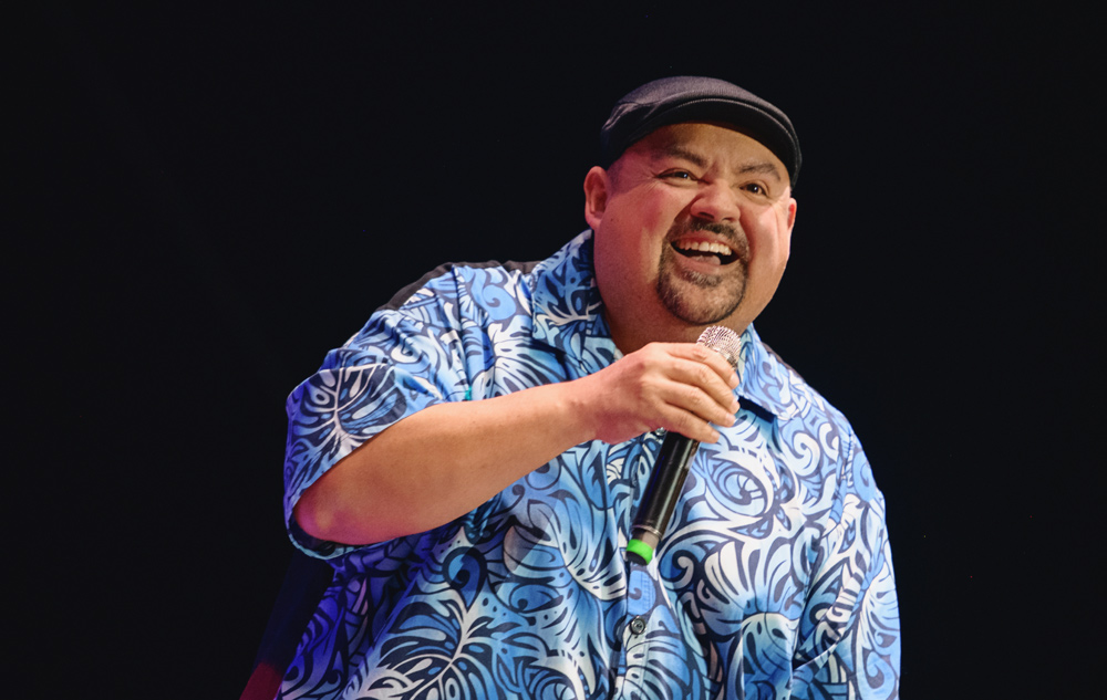 Comedian Gabriel Iglesias laughing into a microphone on stage wearing a blue patterned shirt and flat cap.
