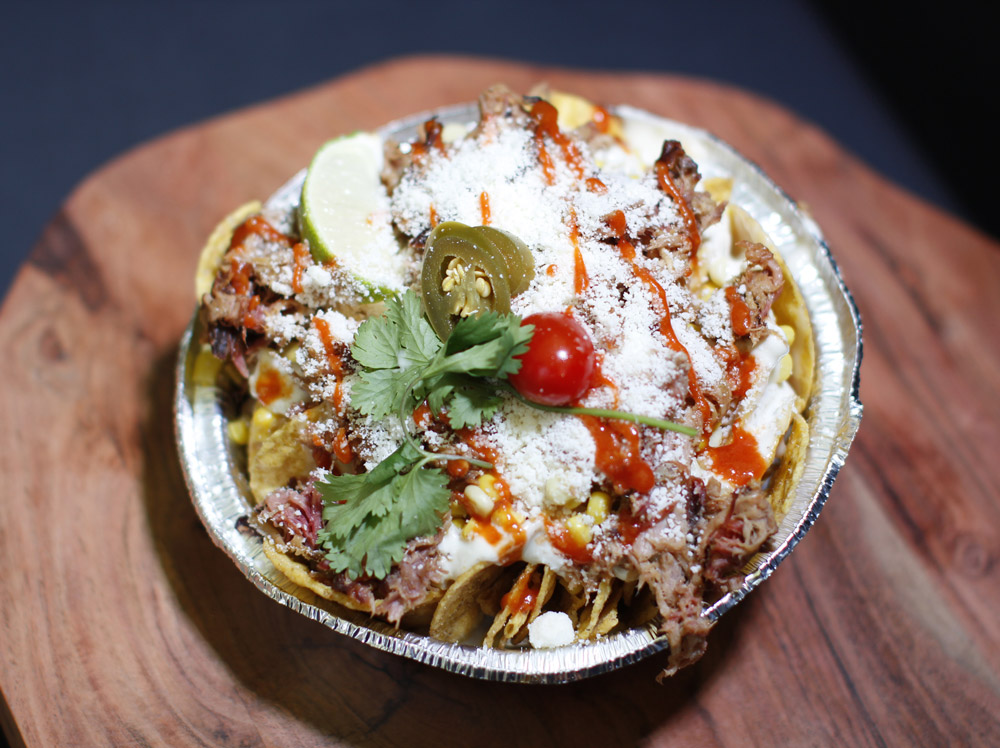 A foil tin loaded with nachos topped with pulled meat, corn, cotija cheese, hot sauce, jalapeño, and cilantro.
