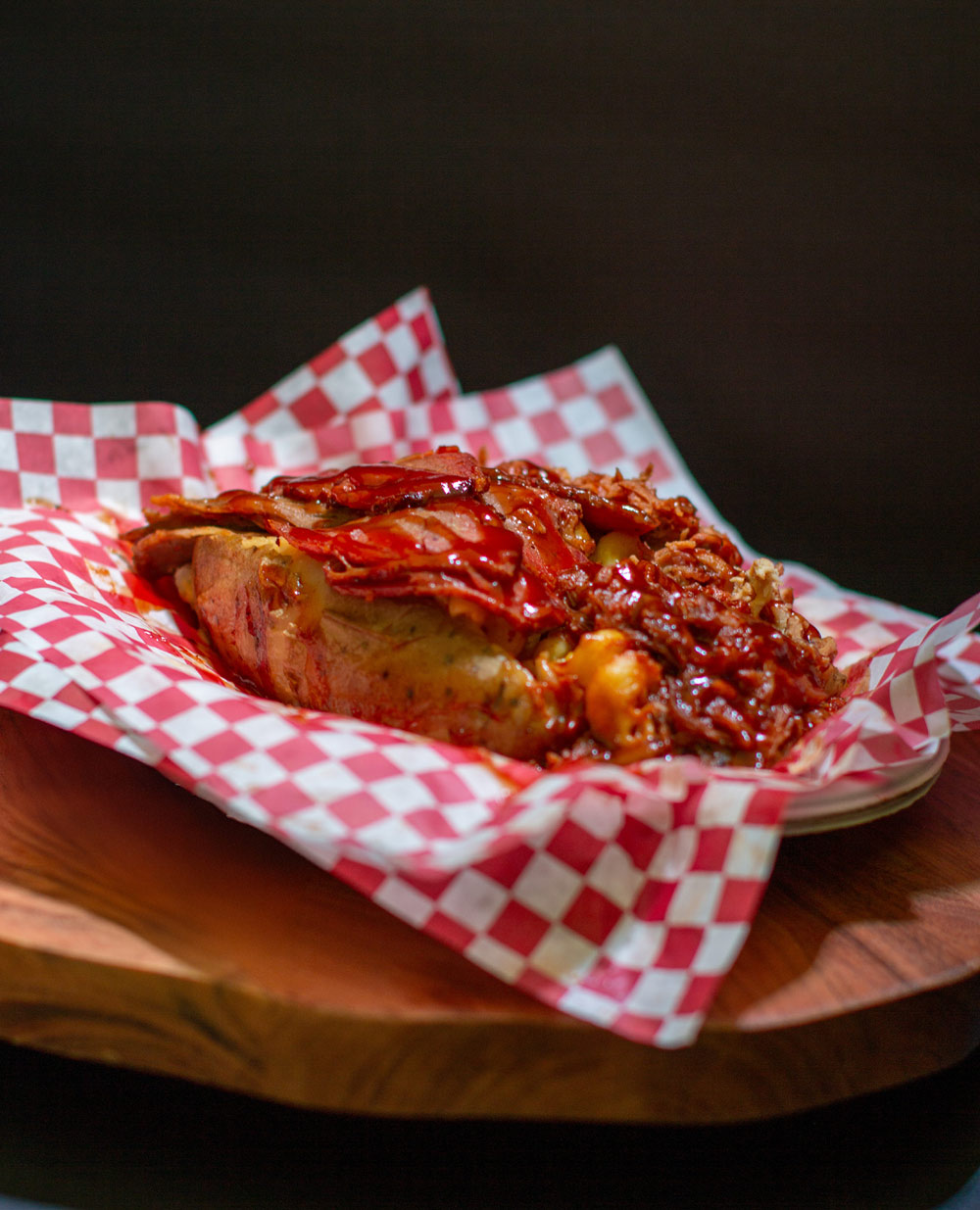 Loaded baked potato piled high with pulled pork, brisket, and barbecue sauce in checkered paper.