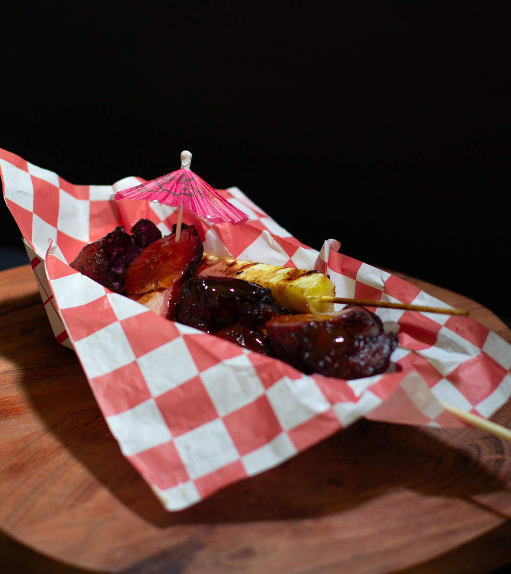 Grilled meat and pineapple skewers garnished with a small pink umbrella in checkered paper.