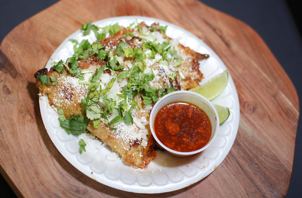 Crispy tacos topped with cilantro, diced onions, and cotija cheese alongside a dipping sauce.