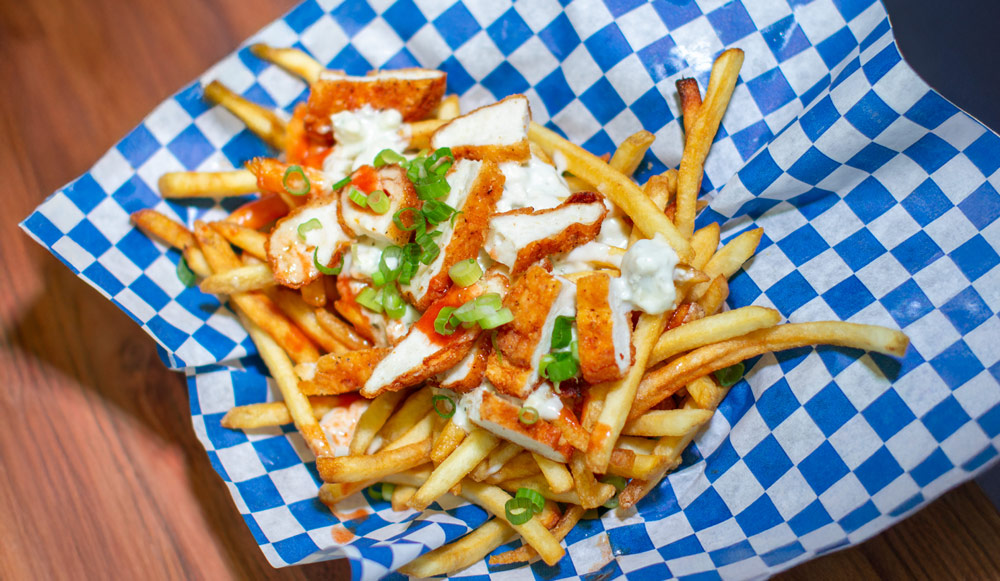 French fries topped with sliced buffalo chicken, green onions, and ranch dressing in checkered paper.