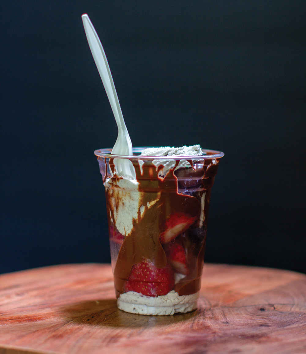 A clear plastic cup filled with fresh strawberries, cream, and drizzled chocolate syrup with a fork inside.