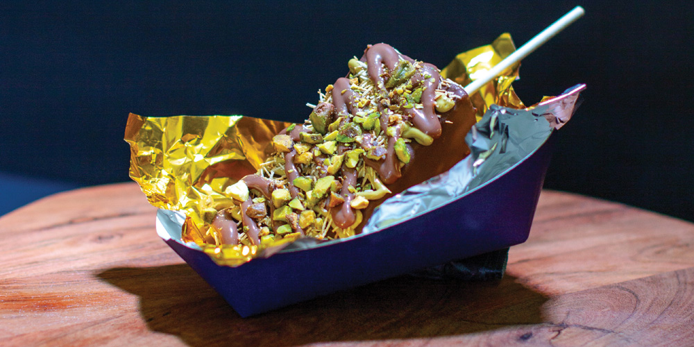 A chocolate-covered dessert on a stick topped with crushed pistachios, served in gold foil inside a paper boat.