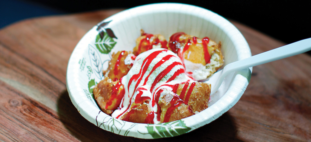 Fried dough bites topped with vanilla ice cream and drizzled red strawberry syrup in a paper bowl.