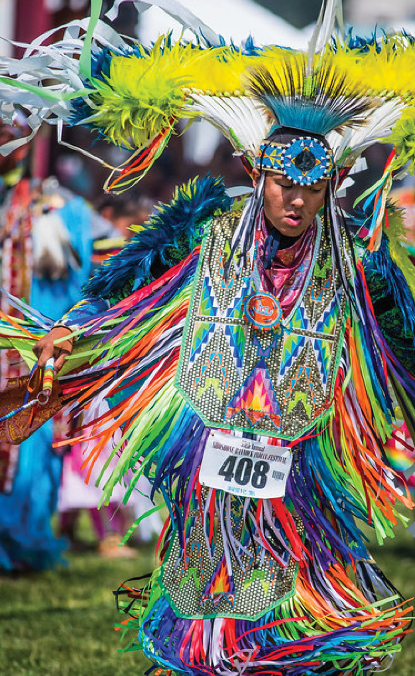 Indigenous dancer in vibrant regalia featuring intricate beadwork, long colorful ribbons, and a large feather headdress, wearing number bib 408 while performing.