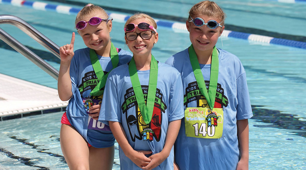Three smiling young girls wearing swim goggles and green medals stand by an outdoor swimming pool in matching blue kids' triathlon shirts.