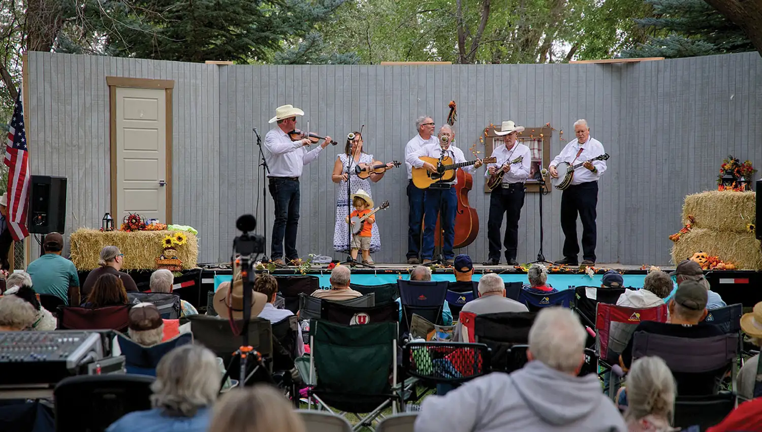A bluegrass band performs on an outdoor stage decorated with hay bales, accompanied by a young child in a cowboy hat, while an audience watches from lawn chairs.
