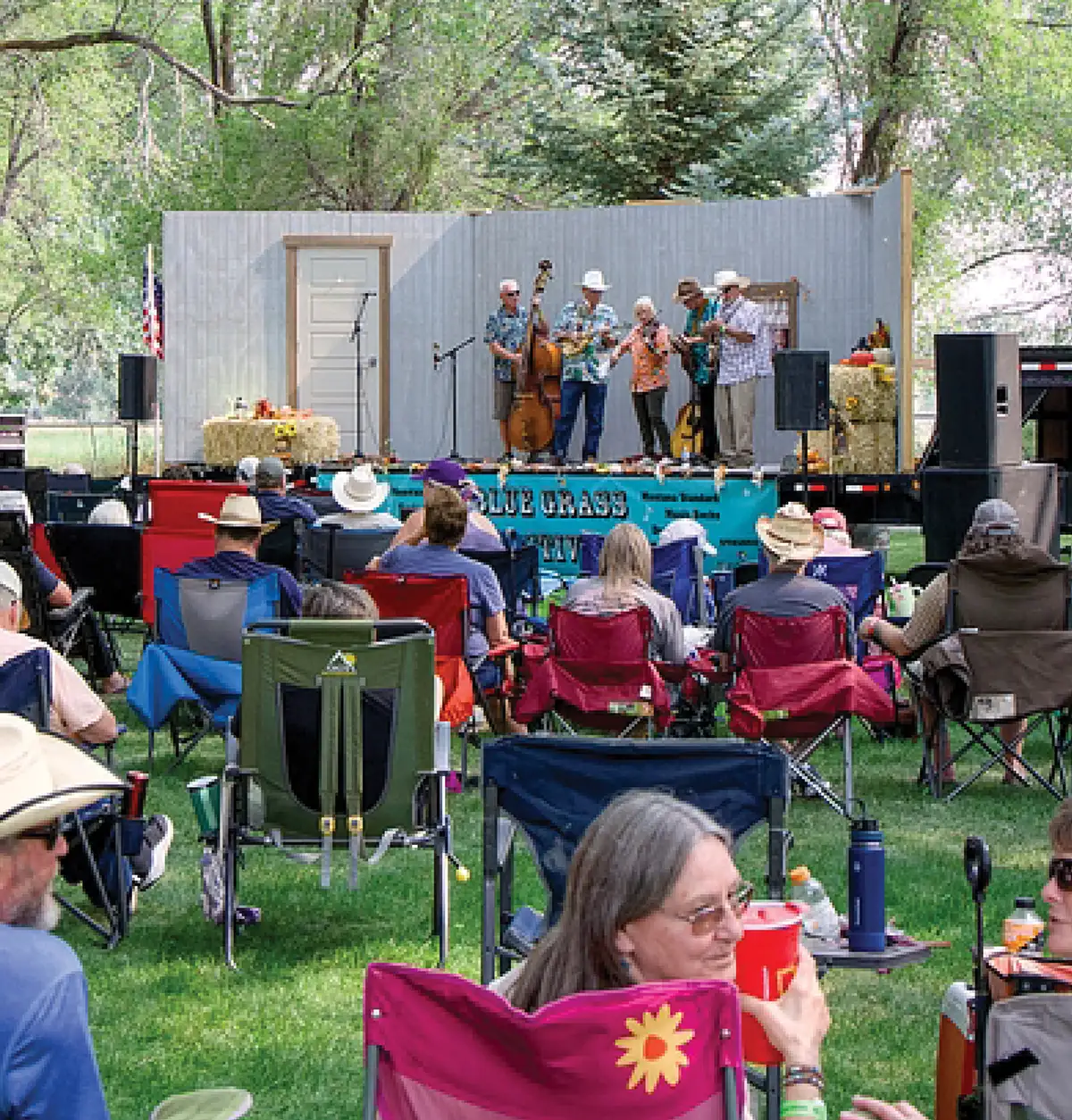 An audience seated in folding lawn chairs watches a live bluegrass band perform on an outdoor stage in a grassy park surrounded by trees.