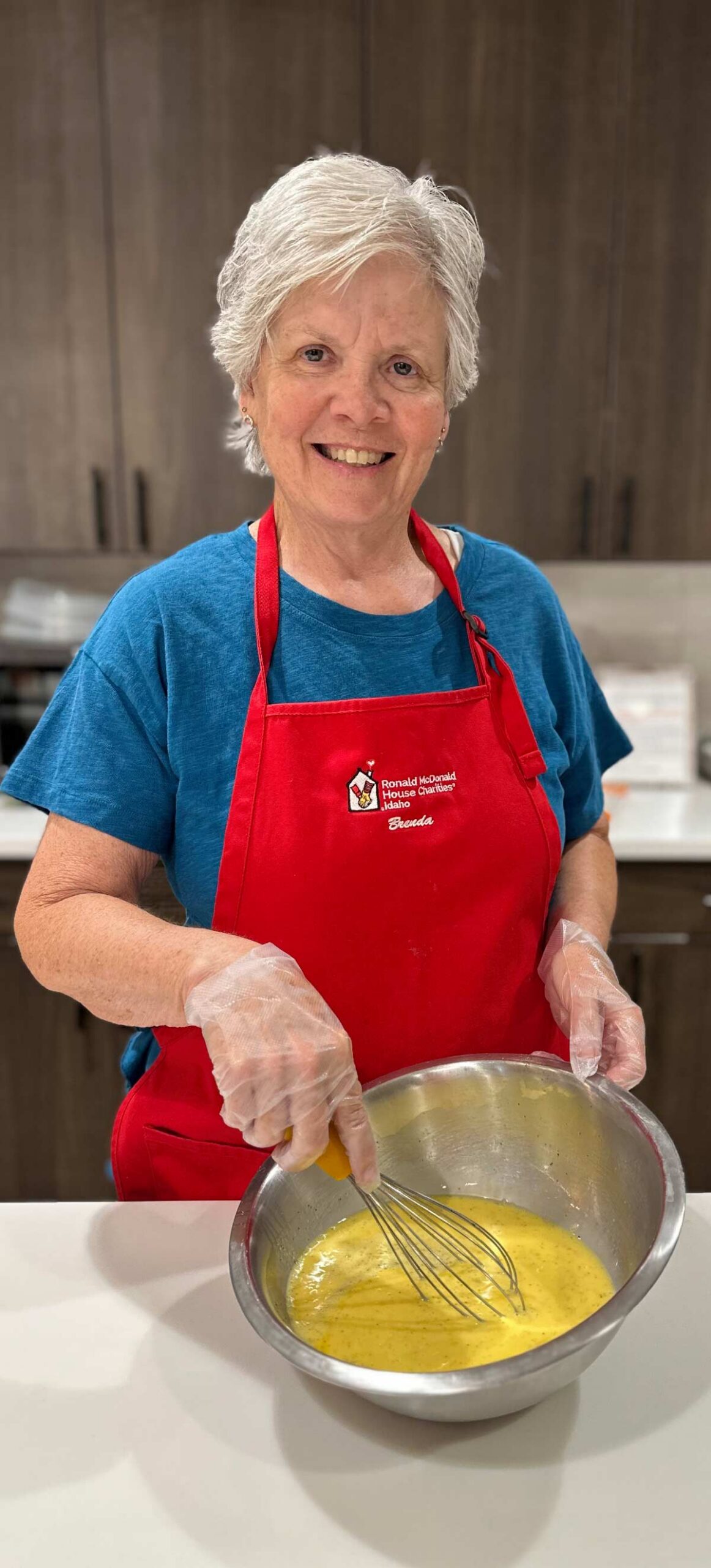 Smiling woman named Brenda in a red Ronald McDonald House Charities apron whisking ingredients in a stainless steel bowl.