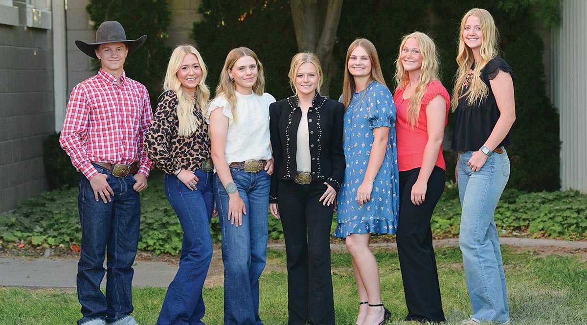 Group portrait of seven smiling young adults in Western-style attire, including jeans, large belt buckles, dresses, and a cowboy hat, posing outdoors.