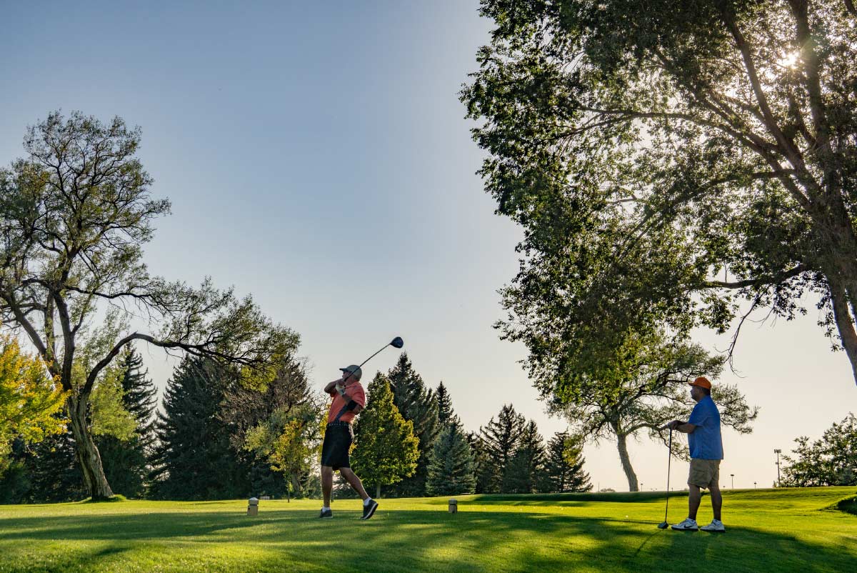Two golfers on a tee box under large shady trees, with one golfer completing a tee shot swing.