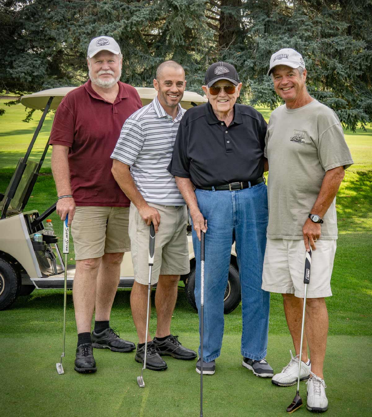Four male golfers smiling and holding putters while posing together on a green near a golf cart.
