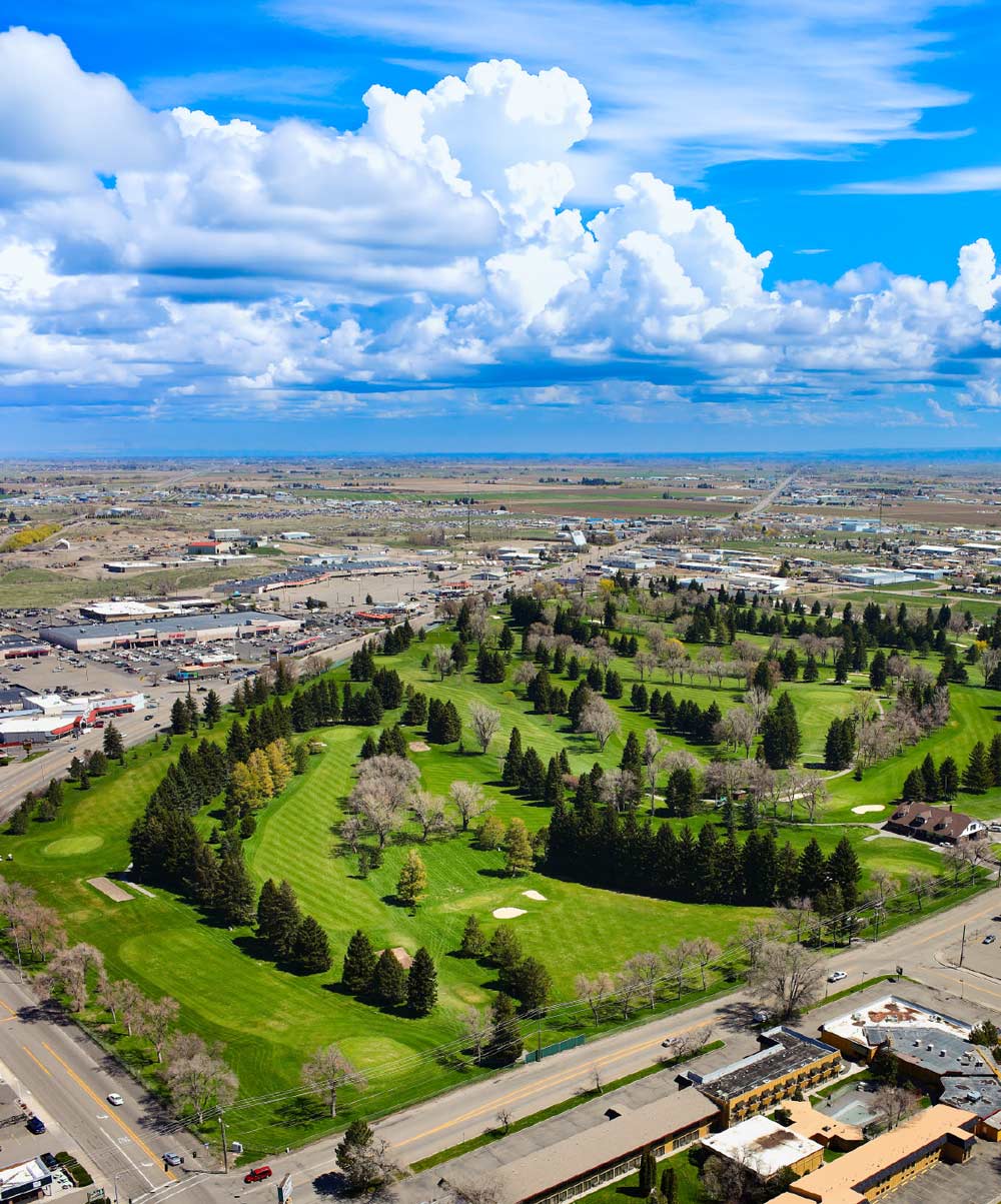 Aerial view of a lush green golf course surrounded by town buildings under a vibrant blue sky with puffy clouds.