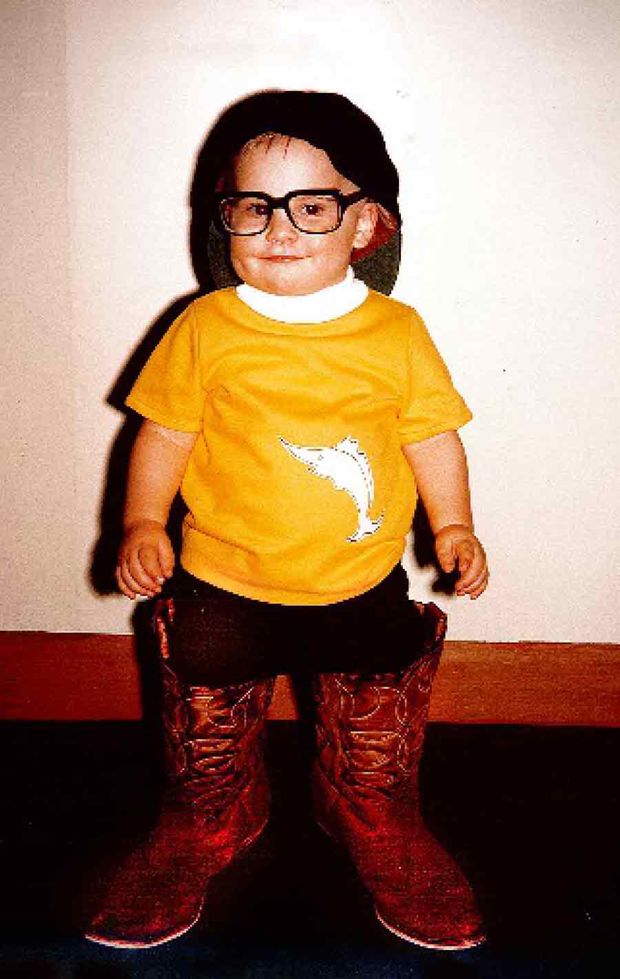 A toddler wearing glasses, a yellow shirt, and oversized cowboy boots.